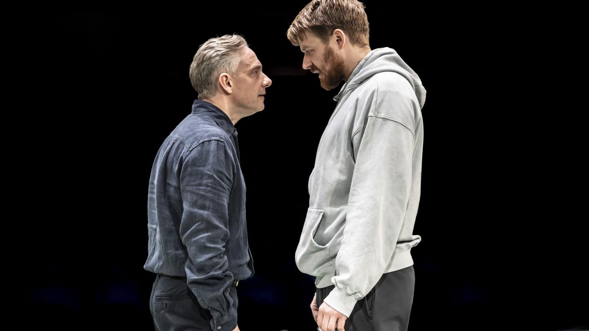 man in black vest and man in gray hoodie staring closely at each other with dark black background