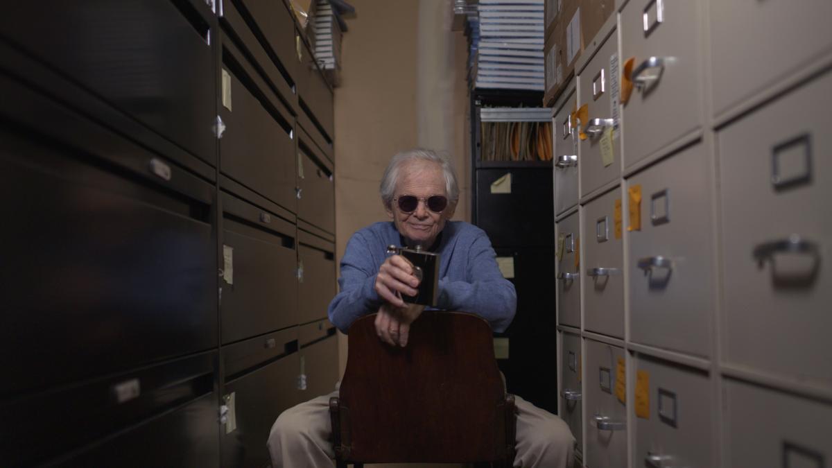 STEVE SCHAPIRO: BEING EVERYWHERE, 2025, man wearing sunglasses straddling backwards chair with file cabinets on either side, holding flask