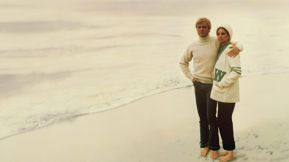 THE WAY WE WERE, 1973, couple wearing sweaters and jeans standing arm and arm on the beach