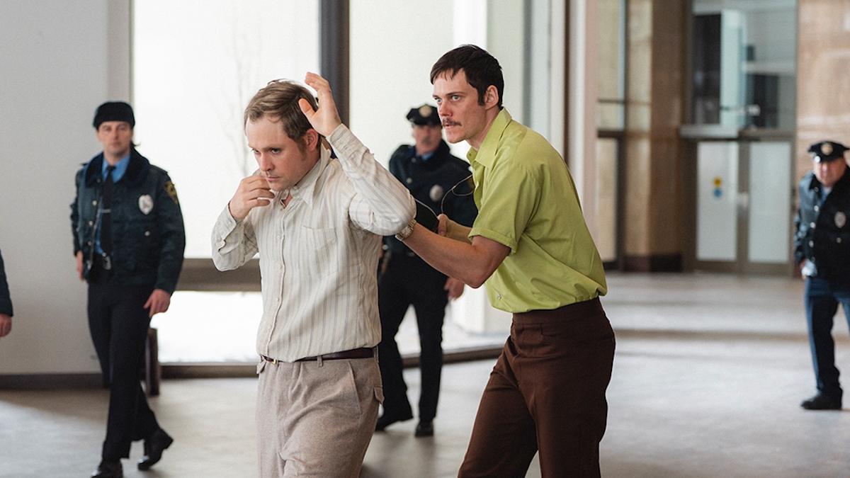 DEAD MAN'S WIRE, 2025, man in green shirt holding gun to other man's back with police officers standing nearby
