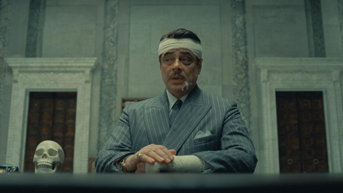 THE PHOENICIAN SHEME, 2025, man in pinstripe suit seated at table with arms crossed and bandage wrapped around his head