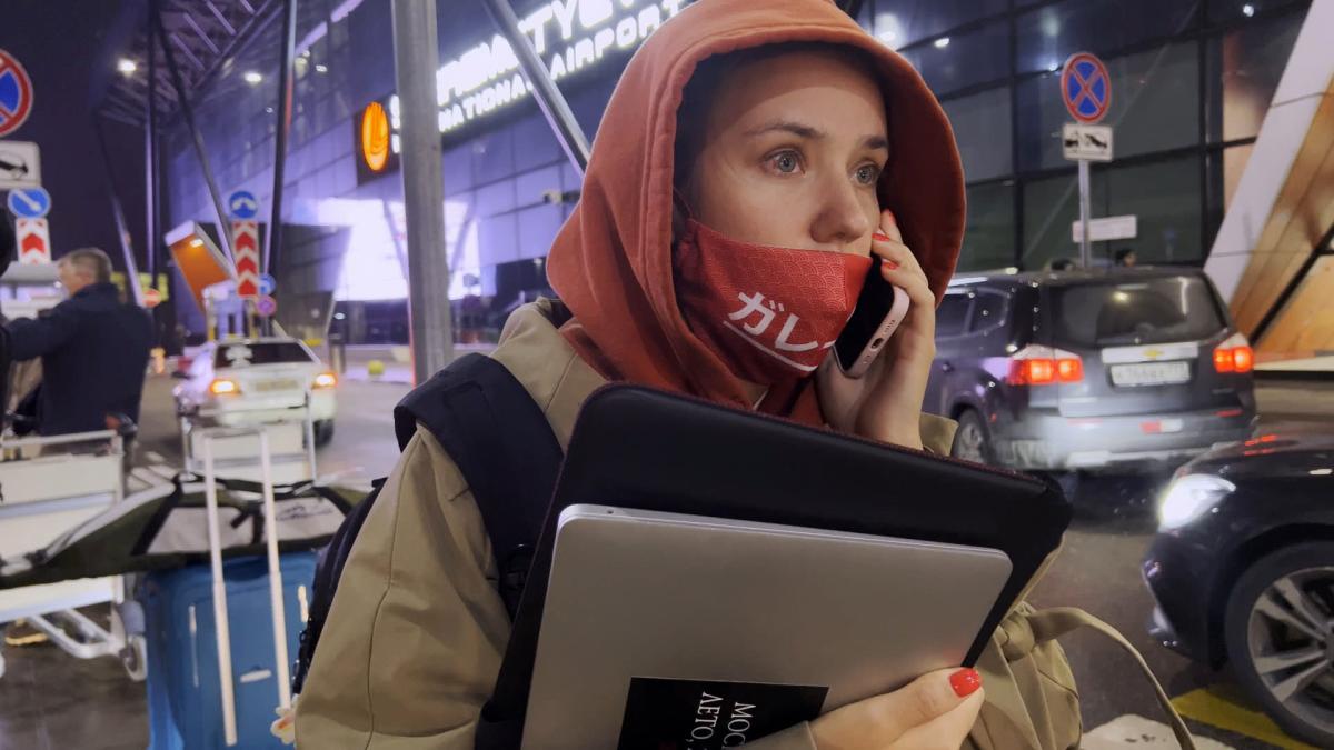 MY UNDESIRABLE FRIENDS: PART I — LAST AIR IN MOSCOW, 2024, girl wearing face mask and hooded sweatshirt talking on phone and walking on street holding laptop