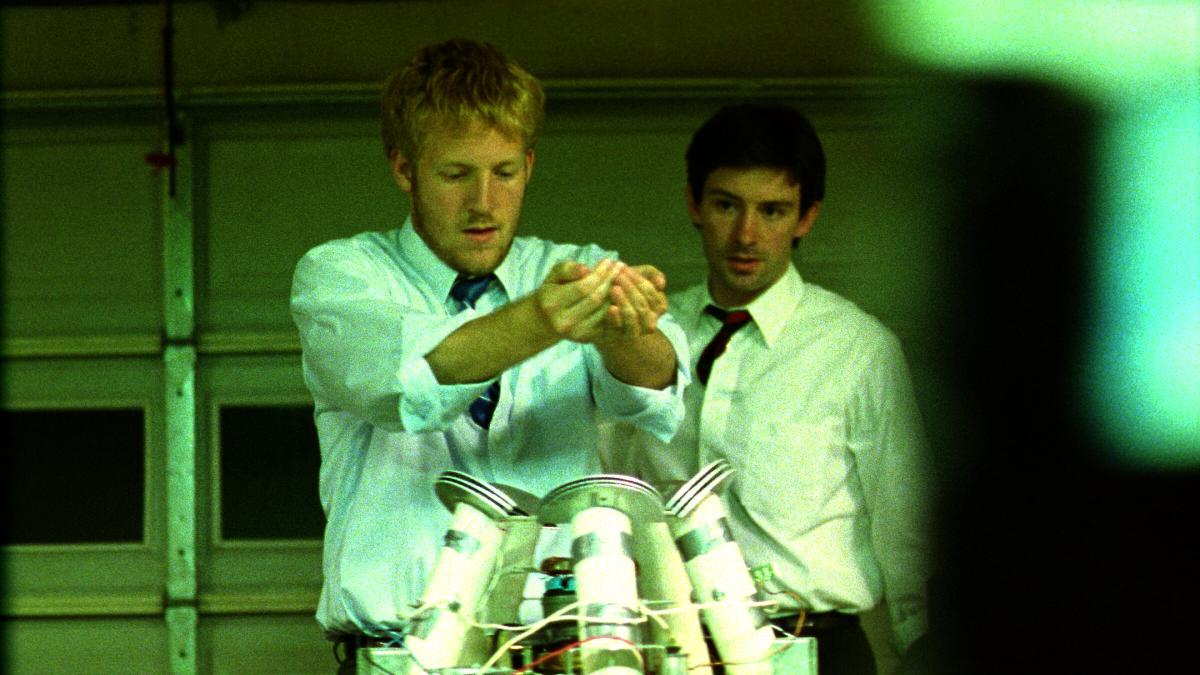 PRIMER, 2004, two men in white shirts and black ties in dimly lit room holding hands over device with wires
