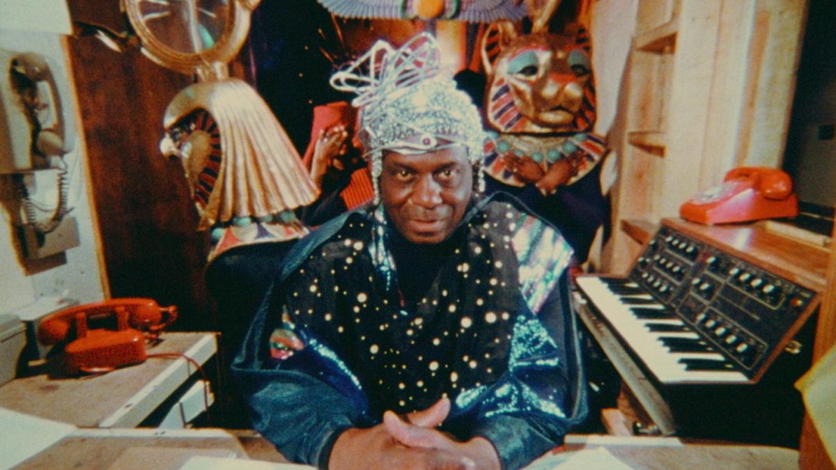 SPACE IS THE PLACE, 1974, Man in ornate elaborate garb and headpiece seated at table with hands folded