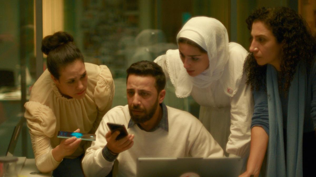 THE VOICE OF HIND RAJAB, 2025, man stiting down on phone at computer with three woman standing around him also on phone with concerned looks on their faces