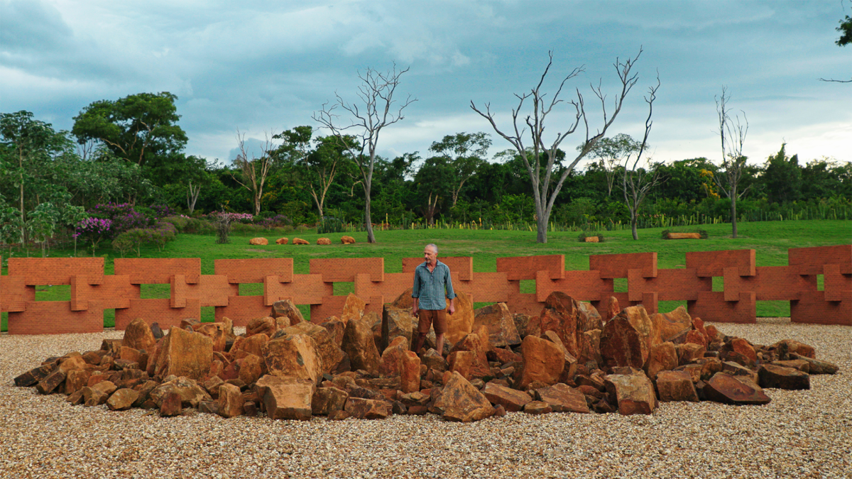 WE THE OTHERS, 2025, man in blue collared shirt standing in pile of large red rocks outside