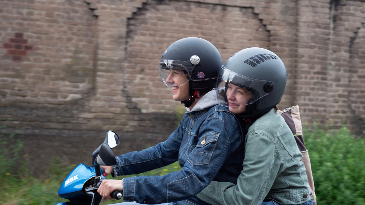 YOUNG MOTHERS, 2025, two people in jackets and helmets riding on moped on street