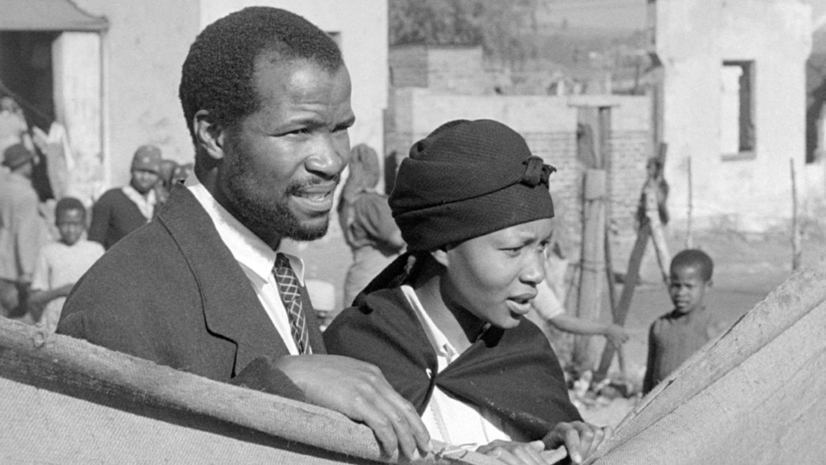 COME BACK, AFRICA, 1959, black and white still of man in suit and woman in scarf peering over wall