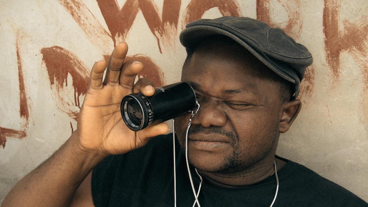 NOLLYWOOD BABYLON, 2008, Black man holding up lens to eye wearing cap