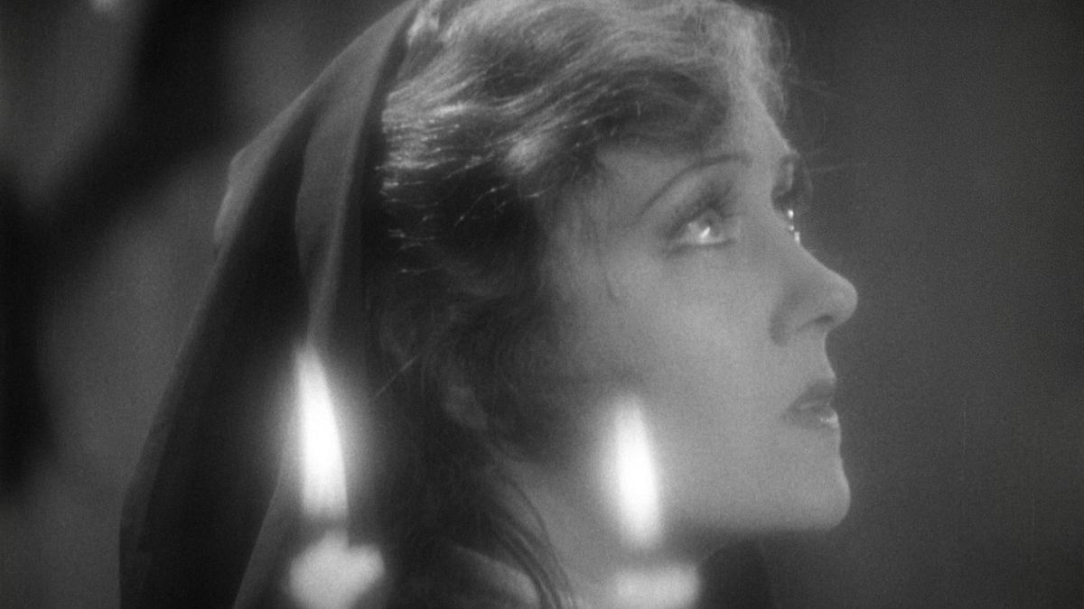 QUEEN KELLY, 1929, black and white still of woman in head scarf looking upwards with candles lit in front of her