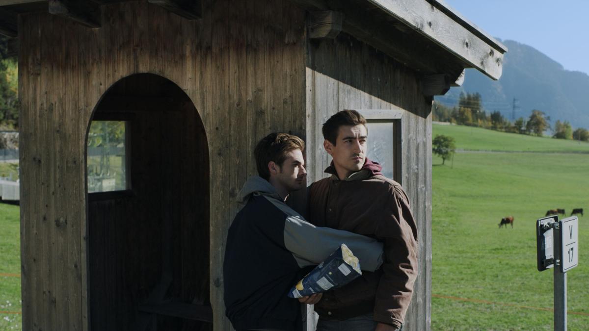 THE MAN WITH THE ANSWERS, 2021, two men standing in front of wooden cabin looking out with fields and mountains in background