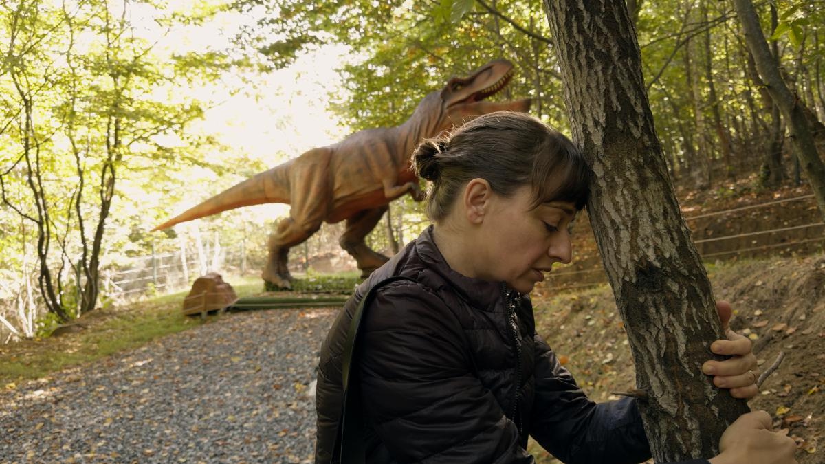 KONTINENTAL '25, 2025, woman leaning over in forest with large t-rex dinosaur behind her