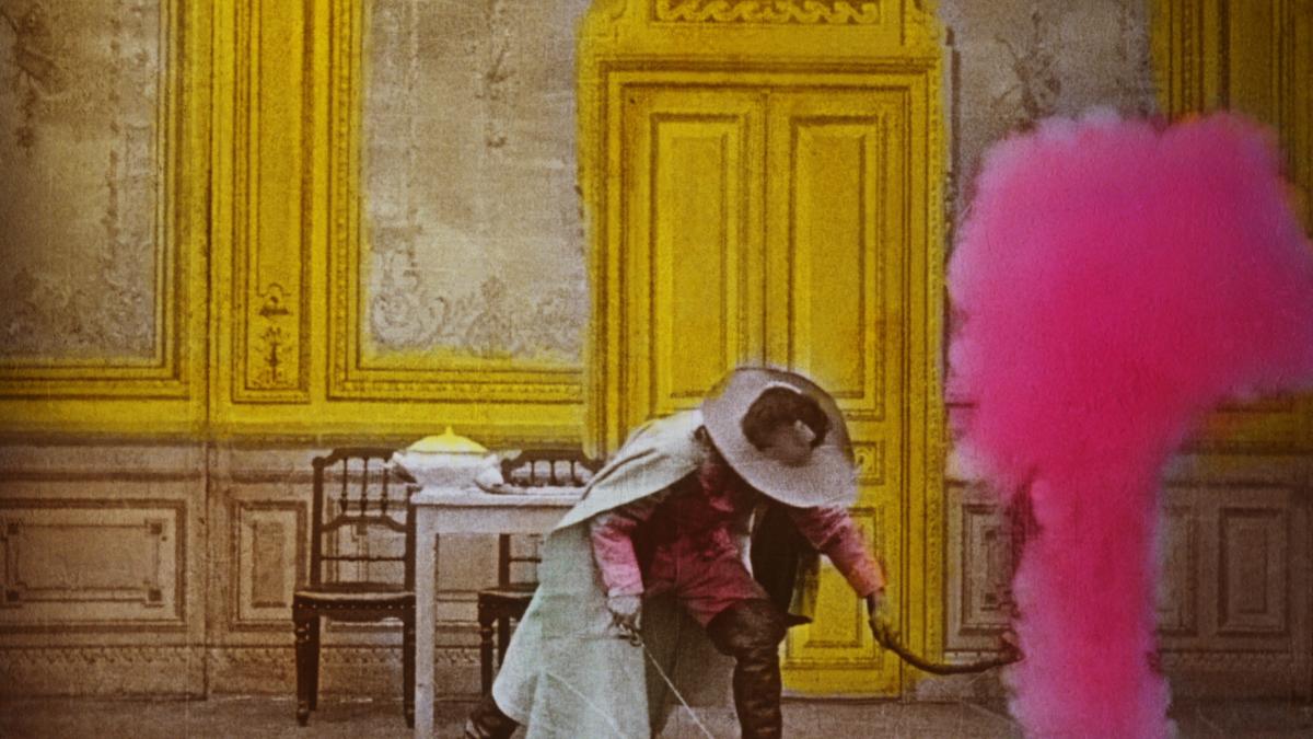 LUMIÈRE, LE CINÉMA!, 2025, technicolor image of man in costume leaning down in room with yellow doors and trim by a plume of pink smoke