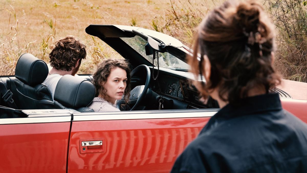 MIROIRS NO. 3, 2025, woman seated in passenger seat of red convertible looking to her right at woman staring at her