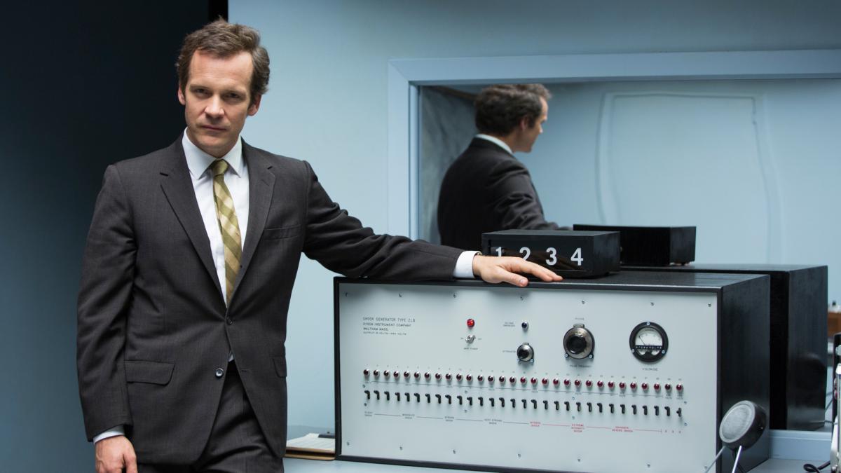 EXPERIMENTER, 2015, man in suit standing with arm resting on large machine