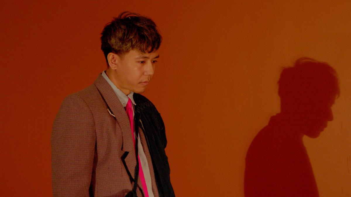 GANGSTERISM, 2025, man wearing jacket standing and looking down with dark orange background