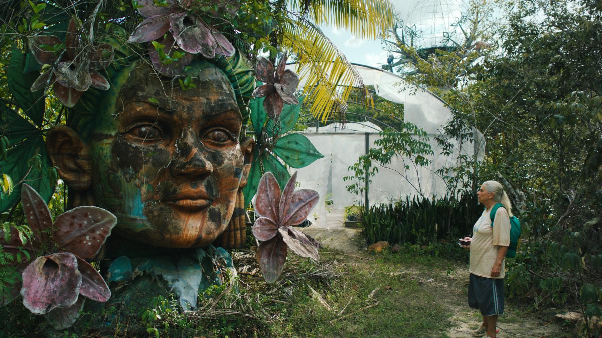 THE BLUE TRAIL, 2025, woman standing in wooded path looking at large bust statue