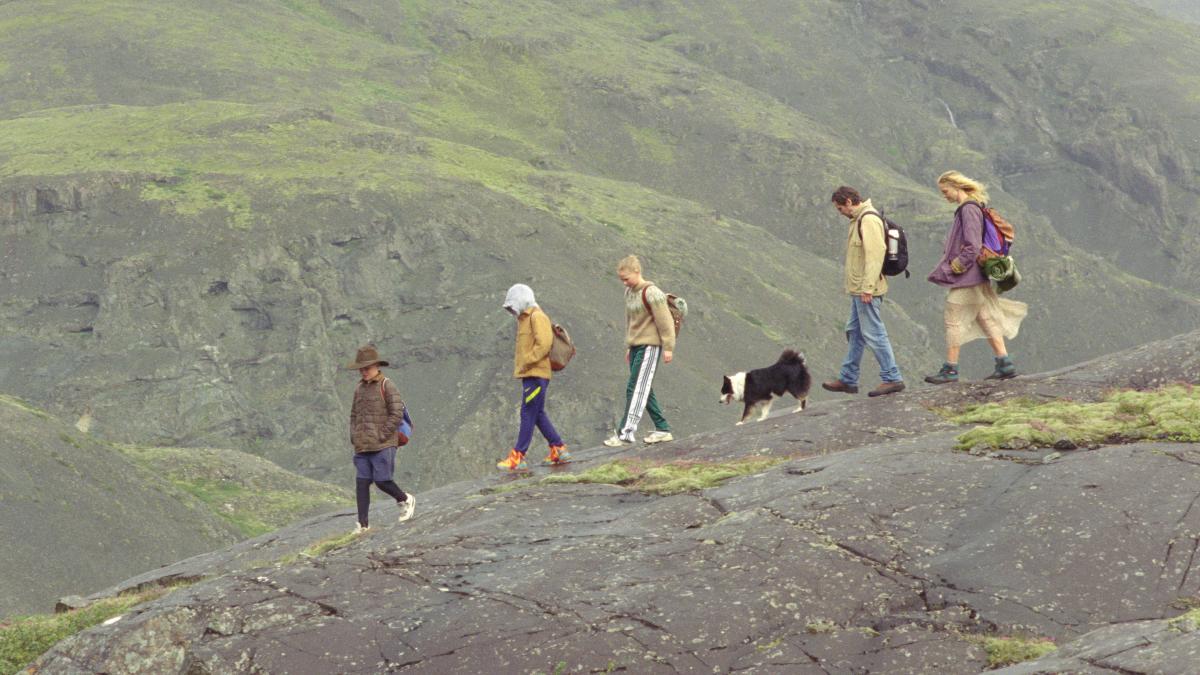 THE LOVE THAT REMAINS, 2025, group of five adults and children with dog walking on rocky terrain in a line