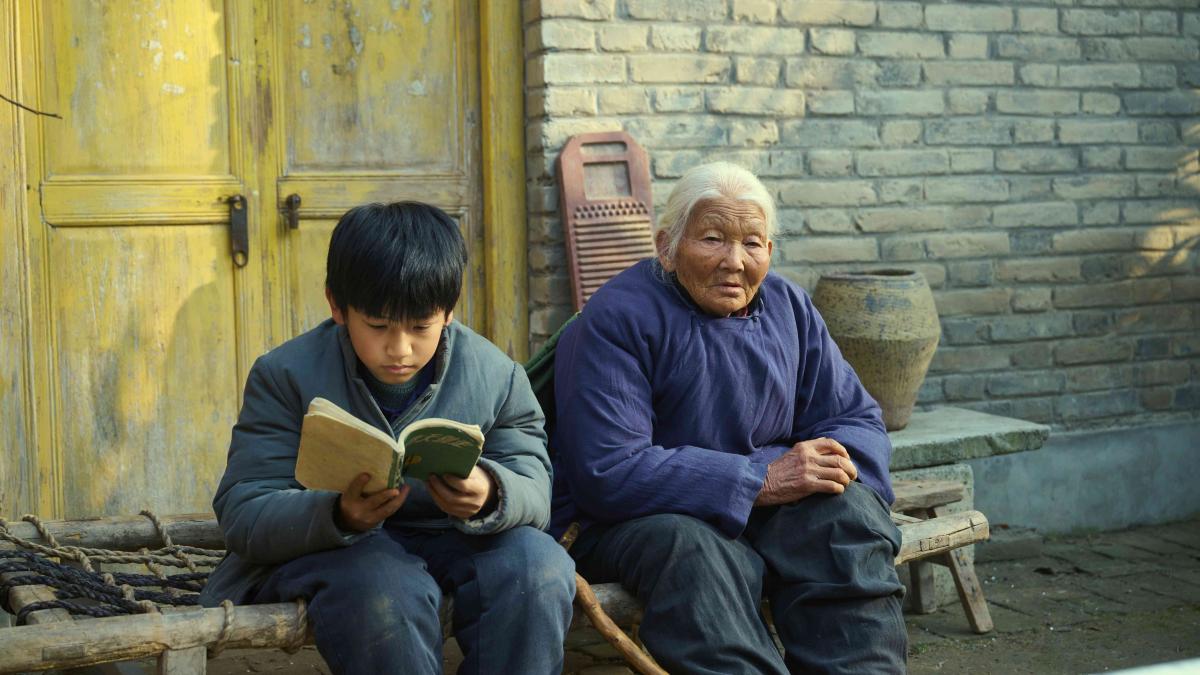 LIVING THE LAND, 2025, elderly woman and a young boy seated on street curb with young boy reading book and a woman softly smiling