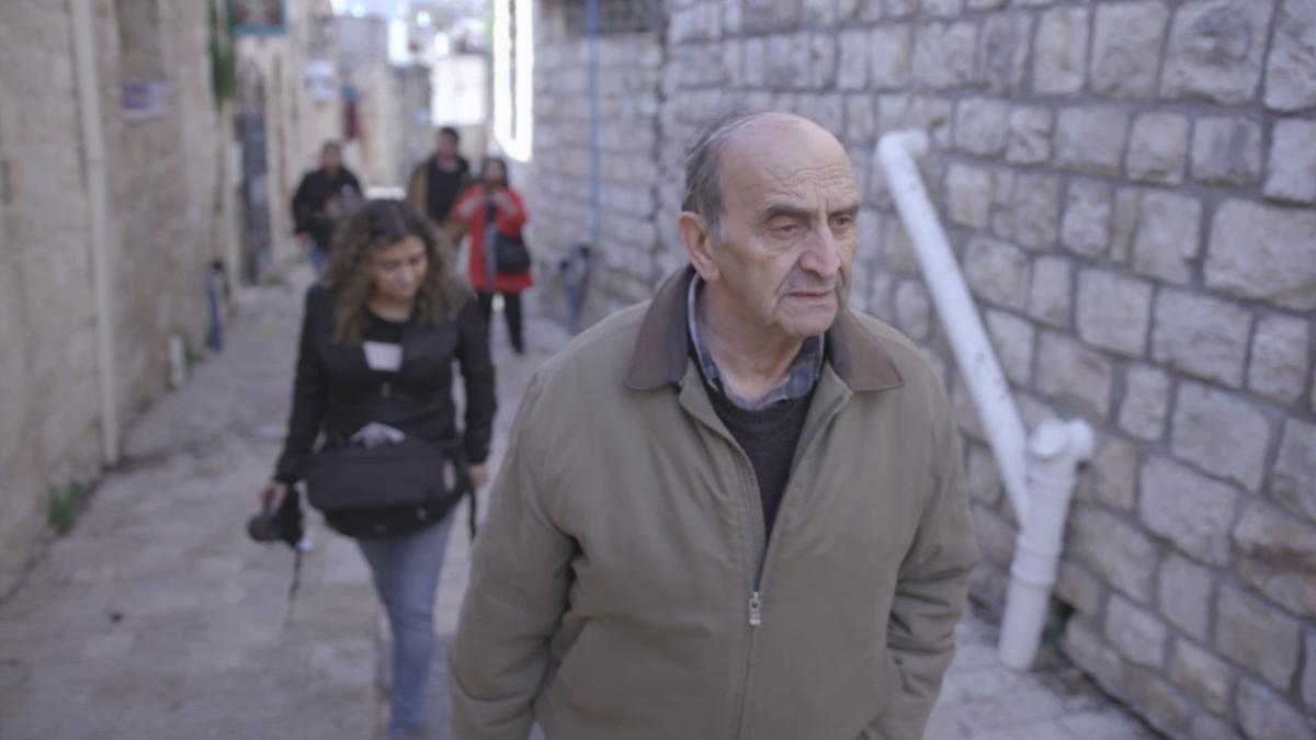 TRACES OF HOME, man in brown jacket walking down alley with a couple of people behind him