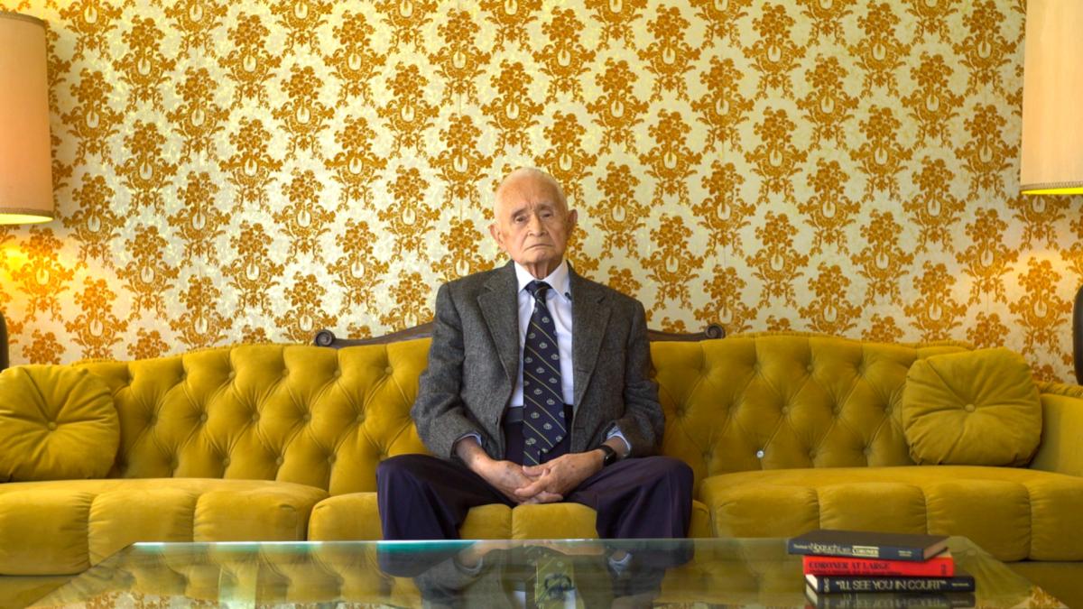 CORONER TO THE STARS, 2025, elderly man in suit seated on yellow sofa with yellow patterned wallpaper behind him