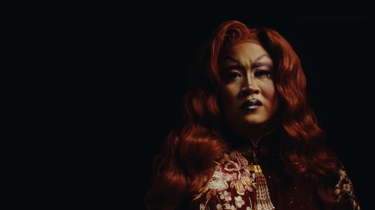 DISSONANCE: AAPI QUEER SHORTS, woman in drag makeup and wig sitting with dim light on her against dark backdrop