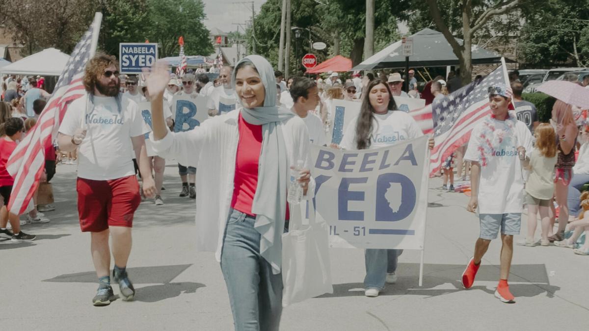 SEAT AT THE TABLE, woman wearing head scarf and holding megaphone walking in parade of people with campaign signs