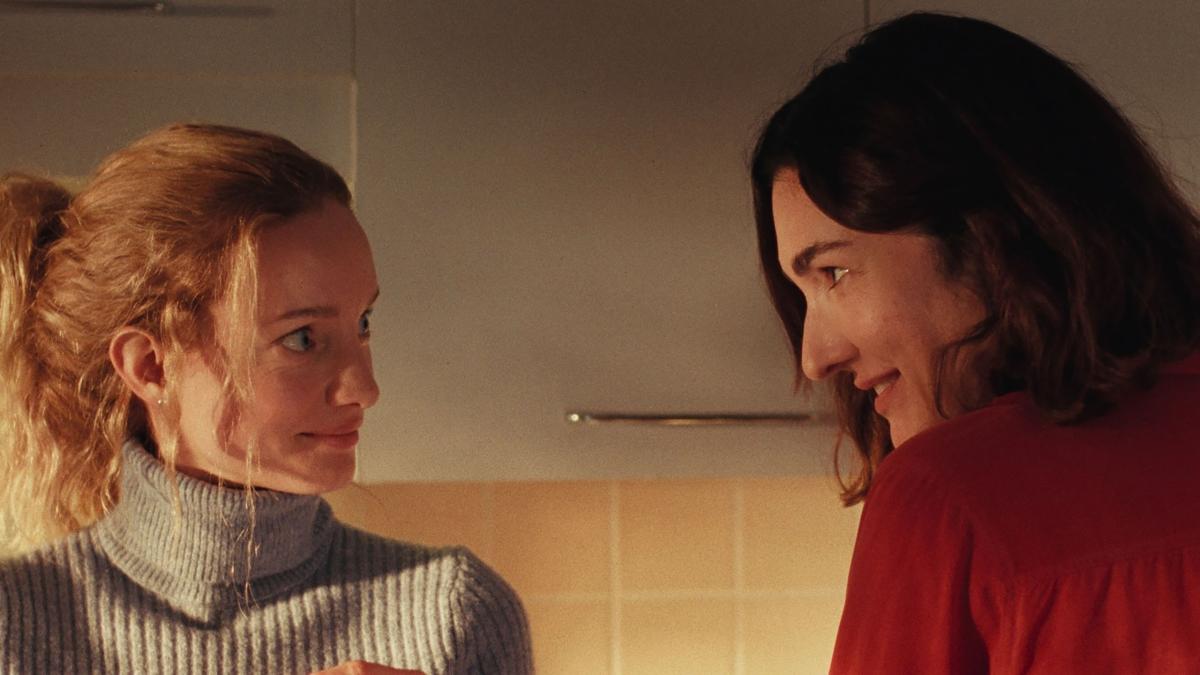 TWO WOMEN, 2025, brunette woman in red sweater looking at blonde woman in grey sweater in room with tiled walls