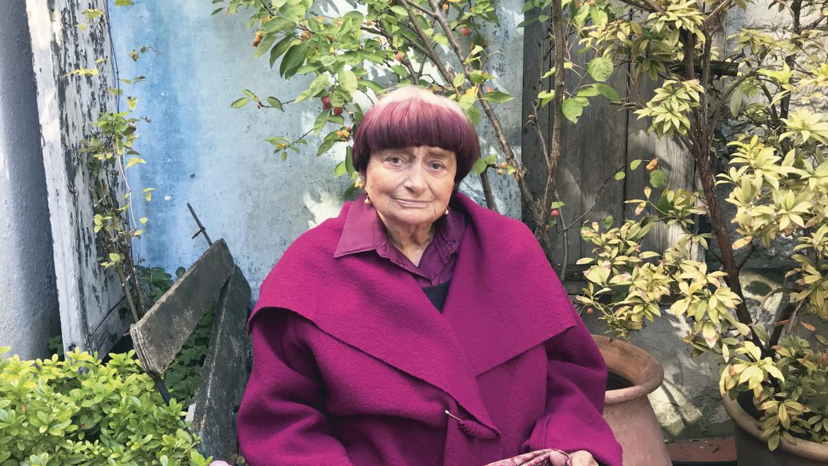 VARDA BY AGNES, 2019, agnes varda in magenta coat seated outside smiling in courtyard
