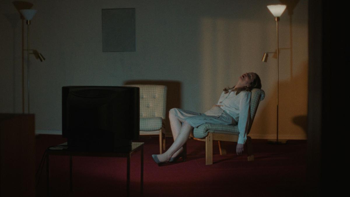 ABOUT A HERO, 2024, girl reclining on chair sleeping with arms hanging by her side in dimly lit living room