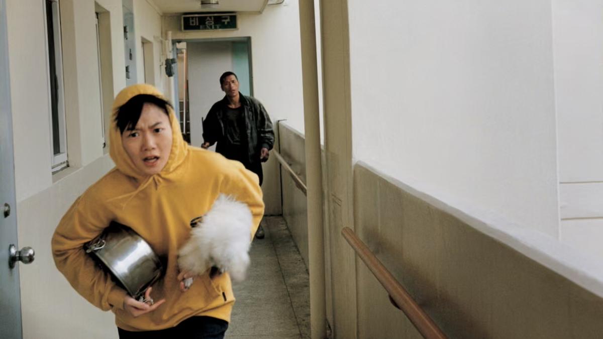 BARKING DOGS NEVER BITE, 2000, woman in yellow sweatshirt running down outdoor corrridor of building with man behind her and holding small white dog and silver pot under her arm