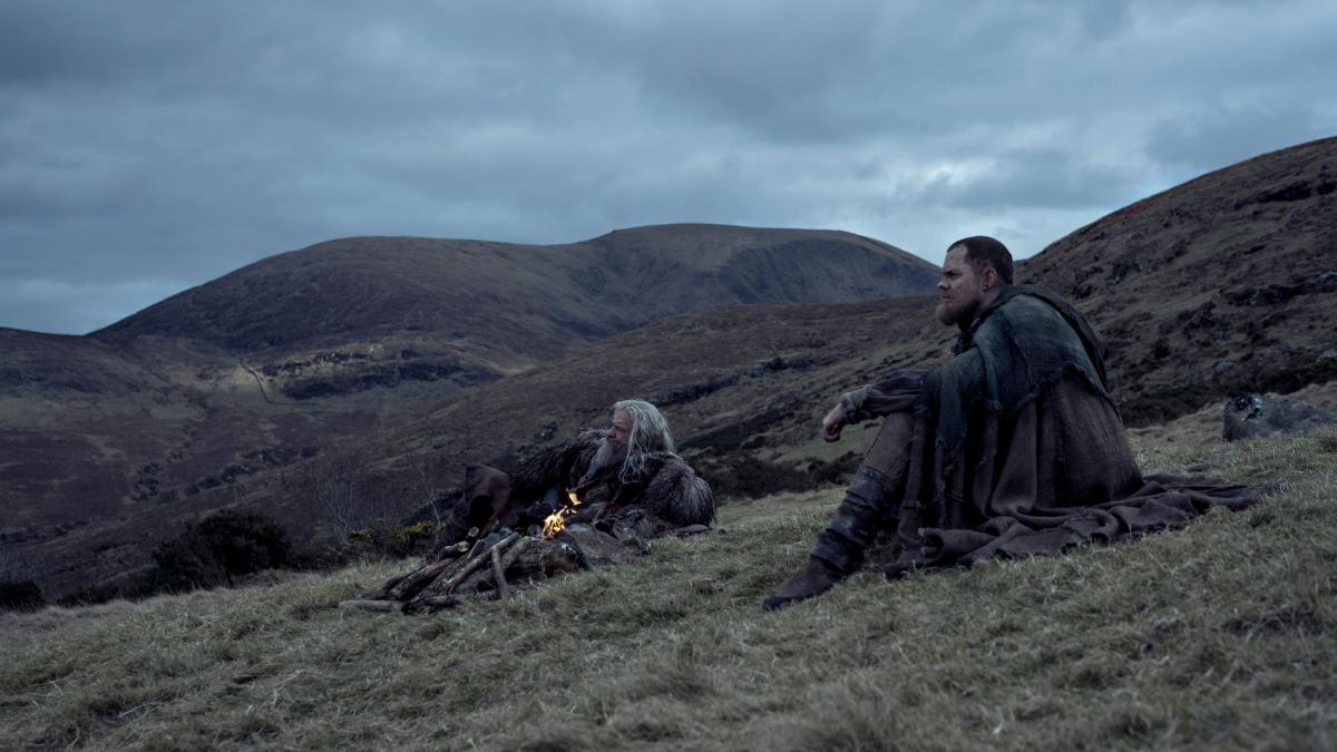 THE DEATH OF ROBIN HOOD, 2026, two men seated on grassy hill looking out into distance with cloudy skies above