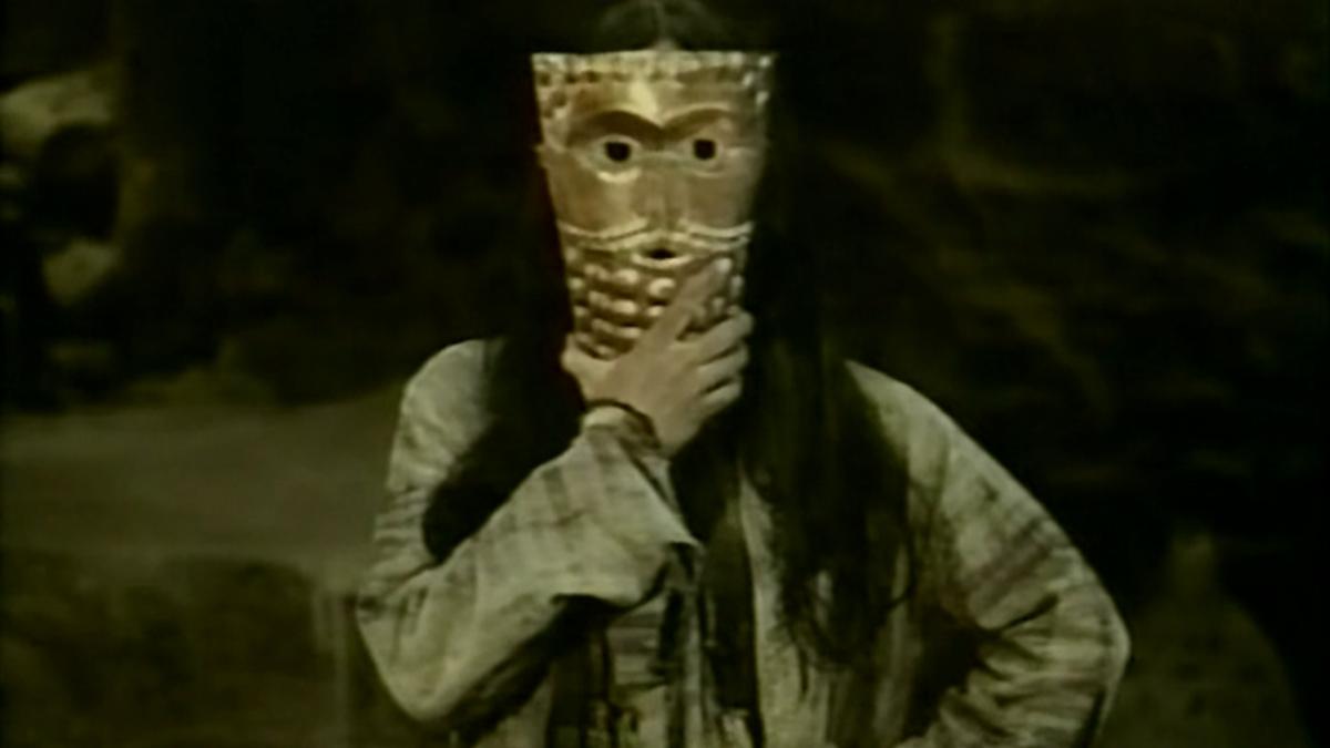 THE DEATH OF YAZDGERD, 1982, person with long hair wearing long sleeves holding up gold mask over face