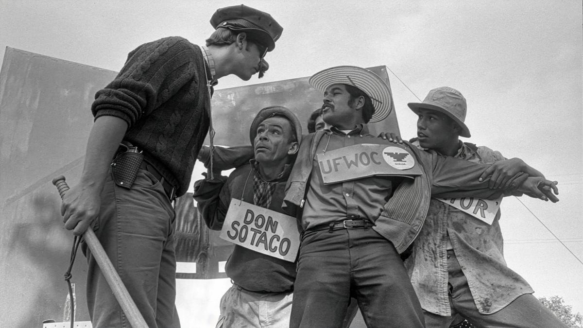 AMERICAN PACHUCO: THE LEGEND OF LUIS VALDEZ, 2026, black and white image taken from below with man in working clothes holding various signs