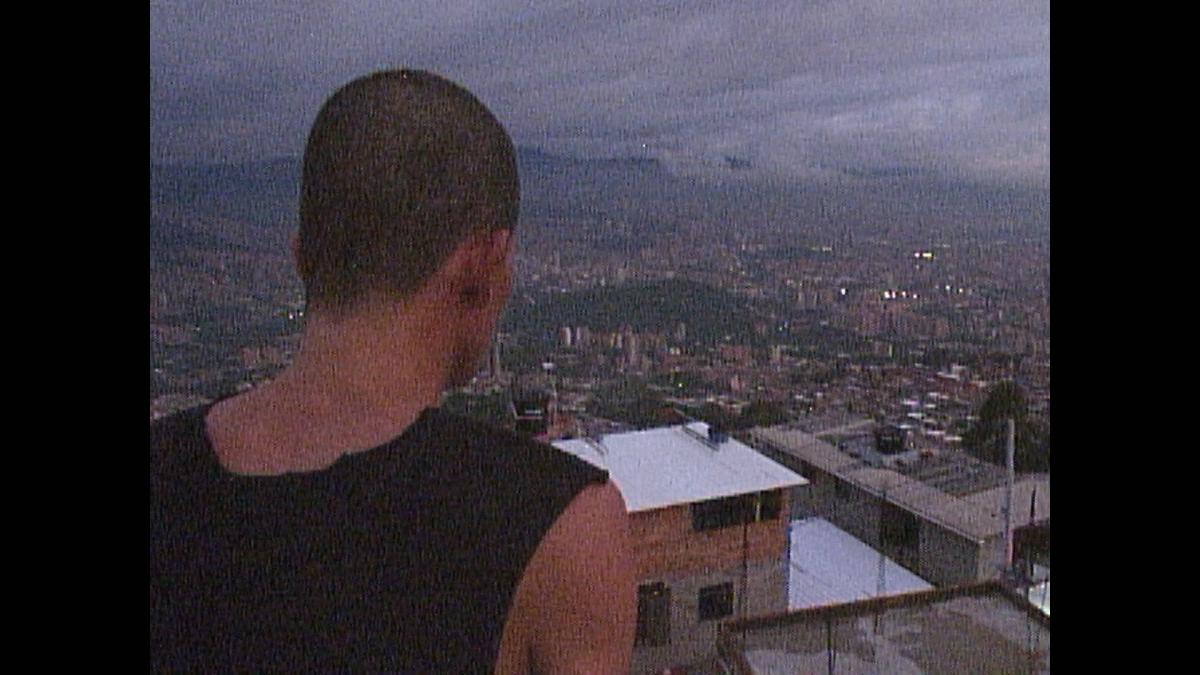 BARRIO TRISTE, 2025, back of man with short hair wearing a tank top overlooking a city with mountains in the distance