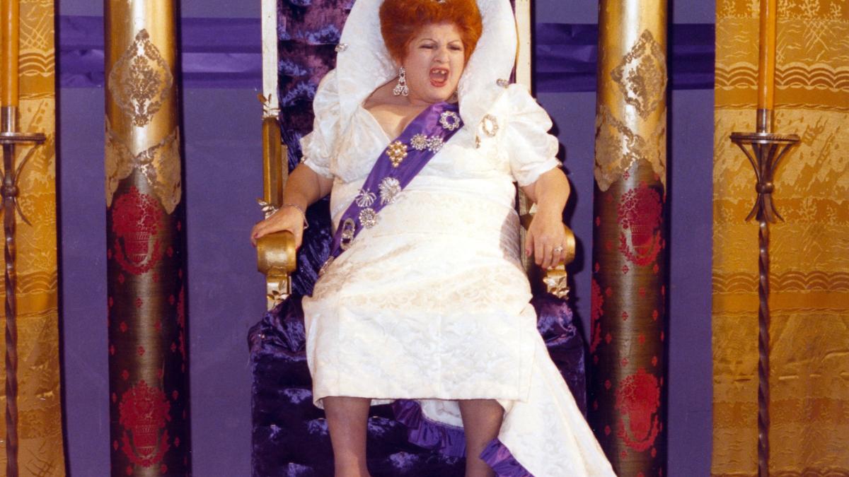 DESPERATE LIVING, 1977, woman in white wedding dress and sash and veil seated on throne with purple and yellow curtains behind