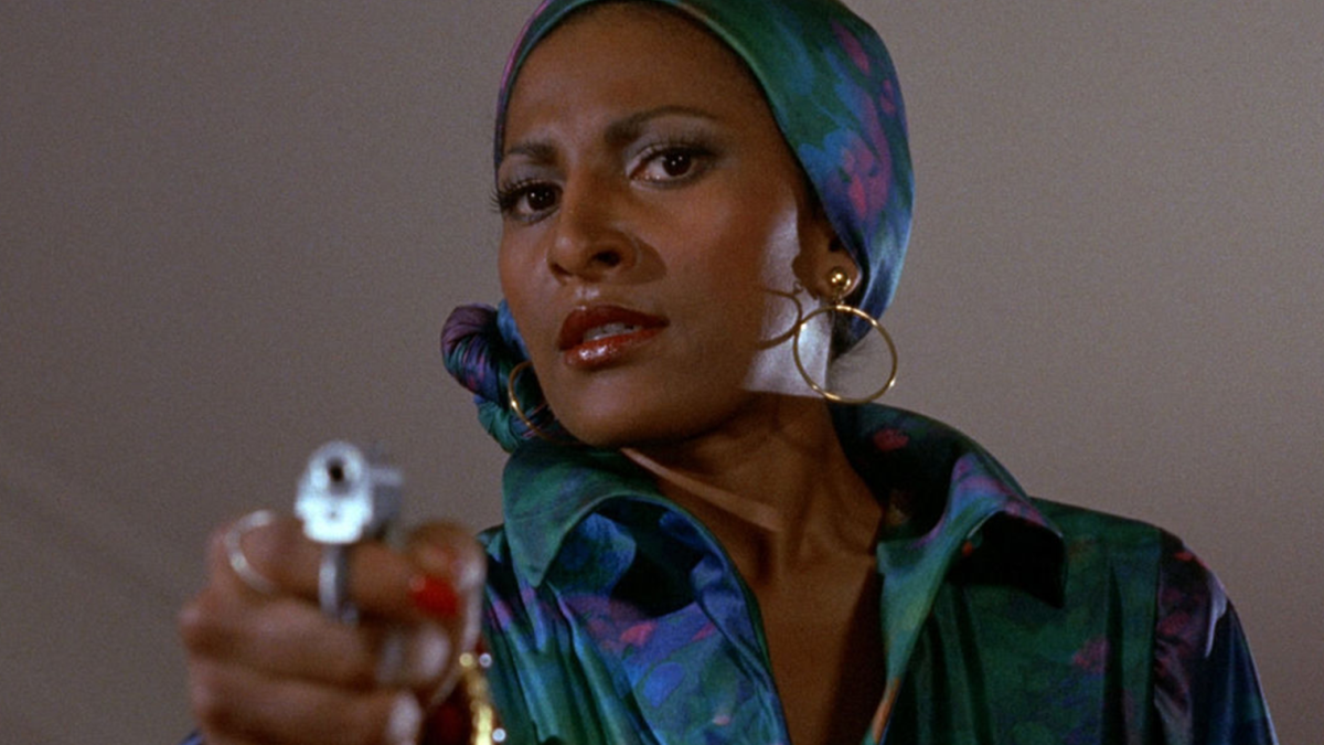 FOXY BROWN, 1974, woman in matching patterned silk shirt and head scarf pointing gun downwards