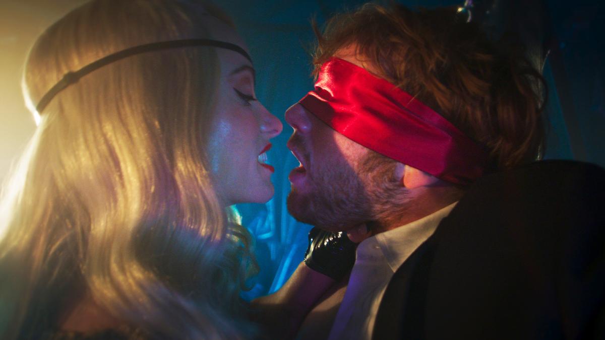 I WANT YOUR SEX, 2025, man wearing red silk blindfold with blonde woman close to his face and holding him