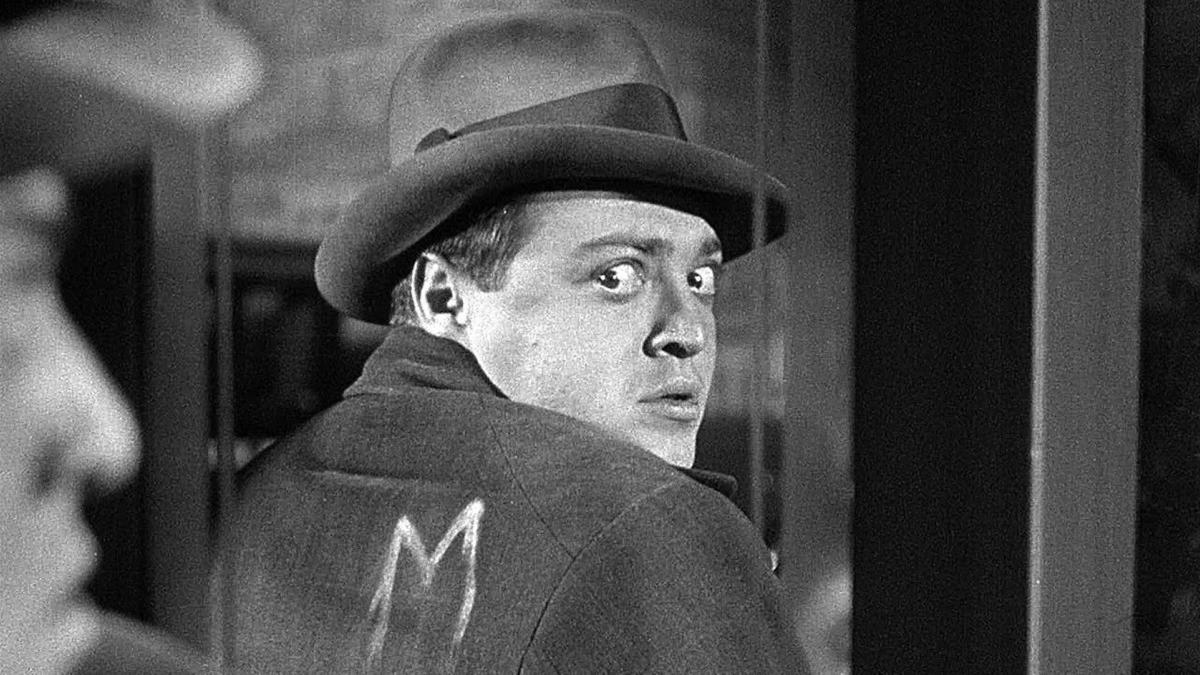 M, 1931, man in jacket and hat turning around in mirror to see the letter M imprinted in his jacket