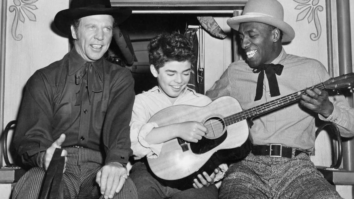 MEET ME AT THE FAIR, 1953, black and white image of young boy holding guitar and smiling with men with western hats seated on either side of him