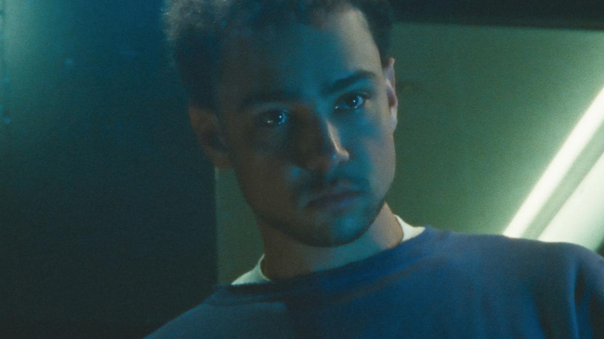 LURKER, 2025, man looking down with fluorescent lights glowing behind him