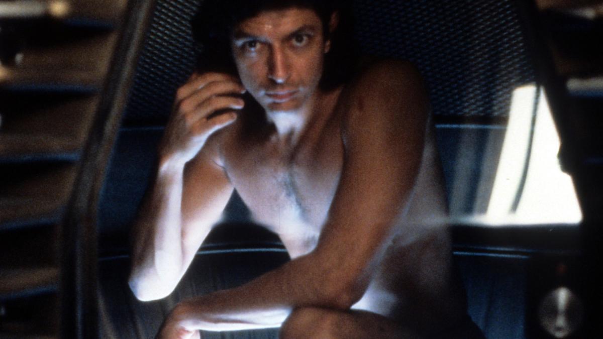 THE FLY, 1986, shirtless man seated in dark holding flashlight in front of him