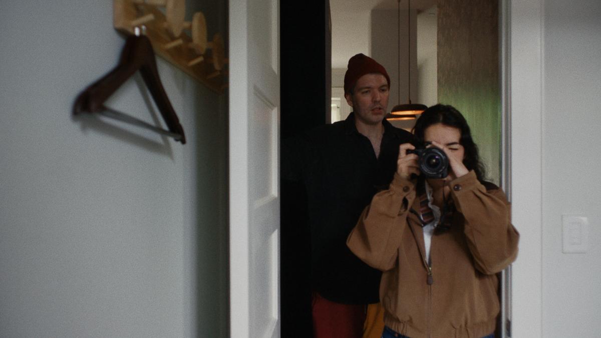 THE SCOUT, 2026, girl wearing brown jacket shooting photo of herself in the mirror with standing behind her in doorway