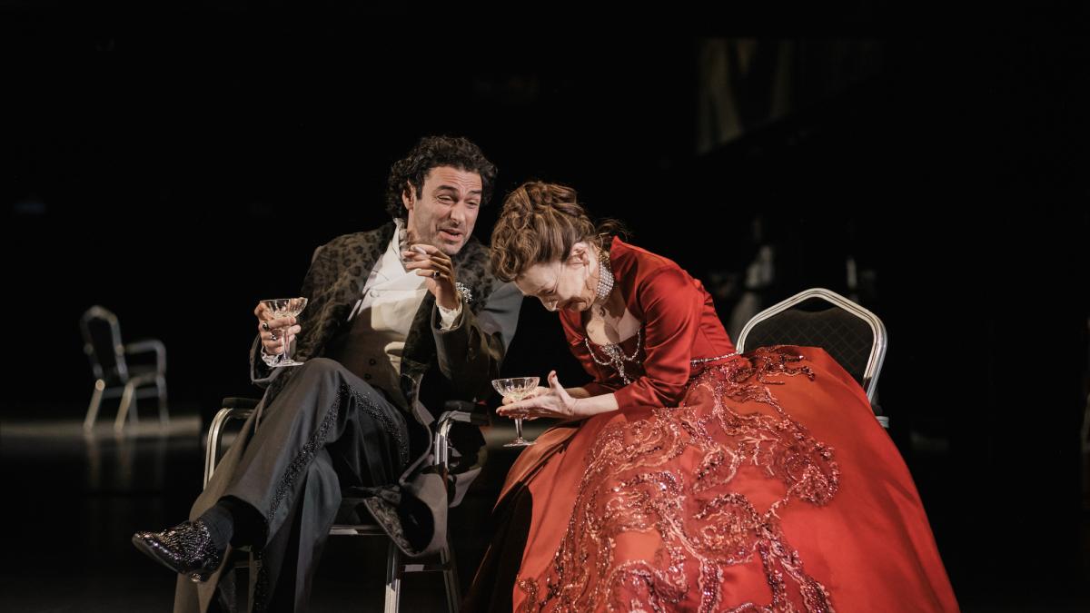 NATIONAL THEATRE LIVE: LES LIAISONS DANGEREUSES, 2026, man in suit and woman in red dress with petticoat seated near each other with woman leaning over