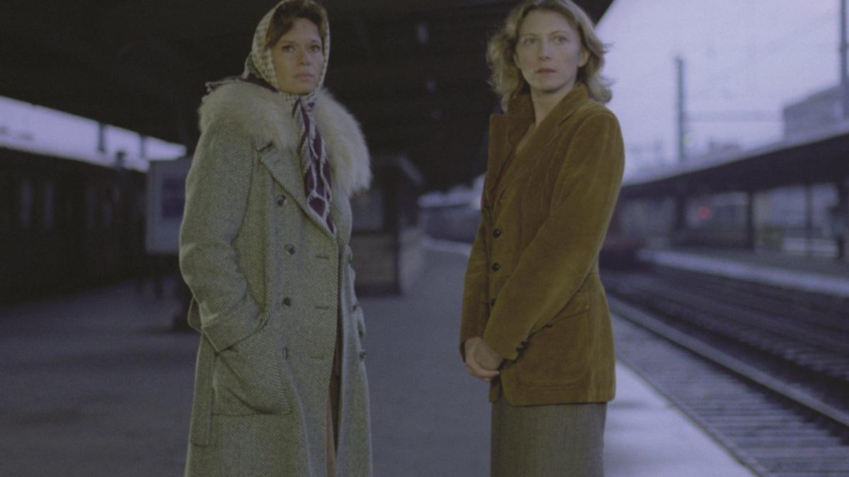 THE MEETINGS OF ANNA, 1978, two women in beige jackets standing on edge of outdoor train platform