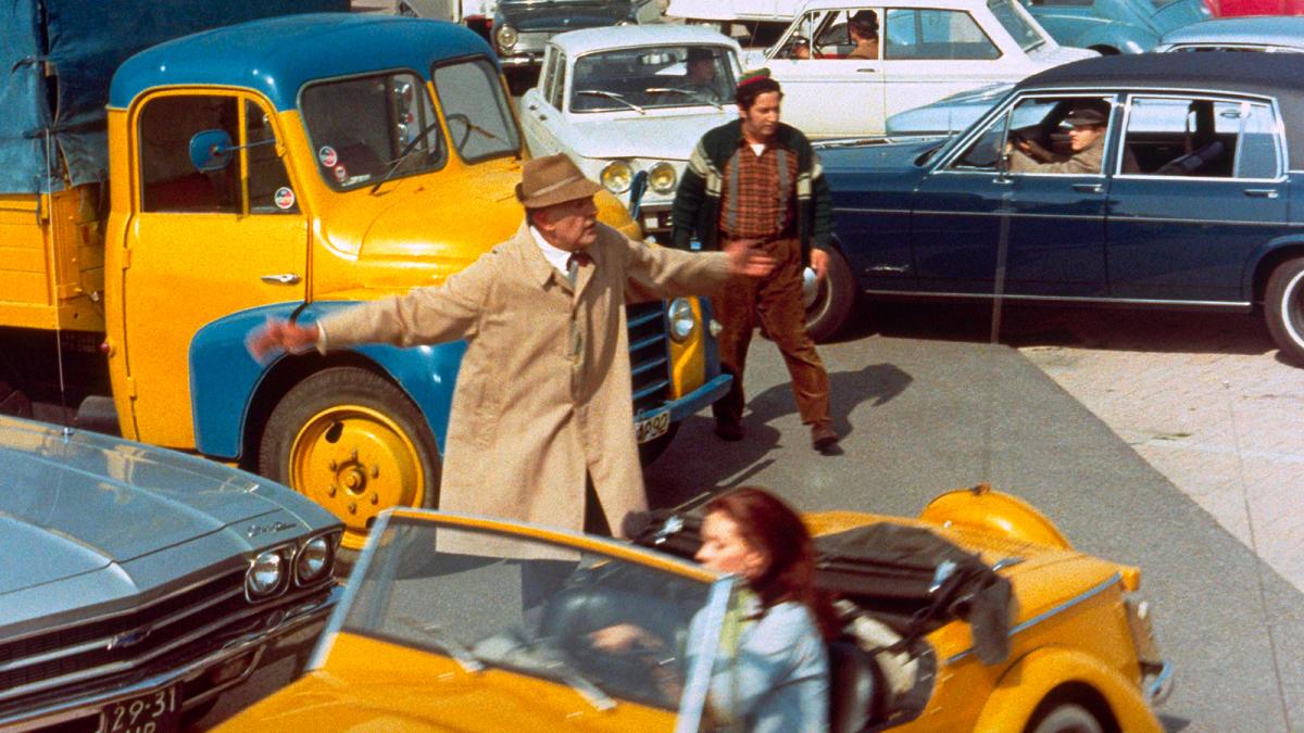 TRAFFIC, 1971, man in trench coat and hat standing in middle of crowded intersection with arms outstretched