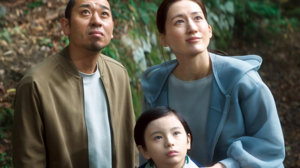SHEEP IN THE BOX, 2026, Japanese father and mother in light jackets looking upwards with mother's arm on young boy's shoulder in front of her