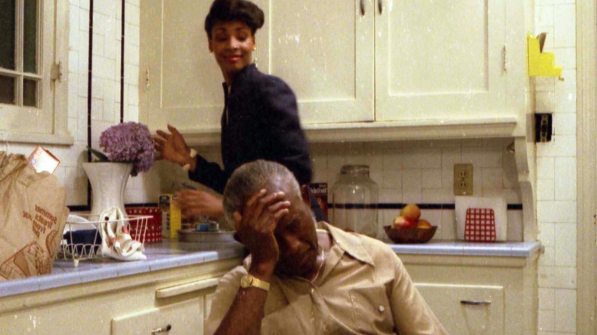 MY BROTHER'S WEDDING, 1983, Black man and woman in kitchen with man on table with head resting on hand and woman standing behind him at the sink