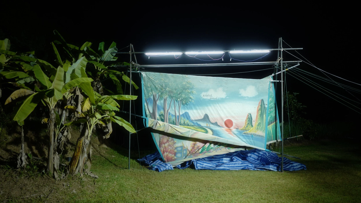 APICHATPONG WEERASETHAKUL SHORTS, image of blue tropical scene on banner hanging from post in dimly lit forest