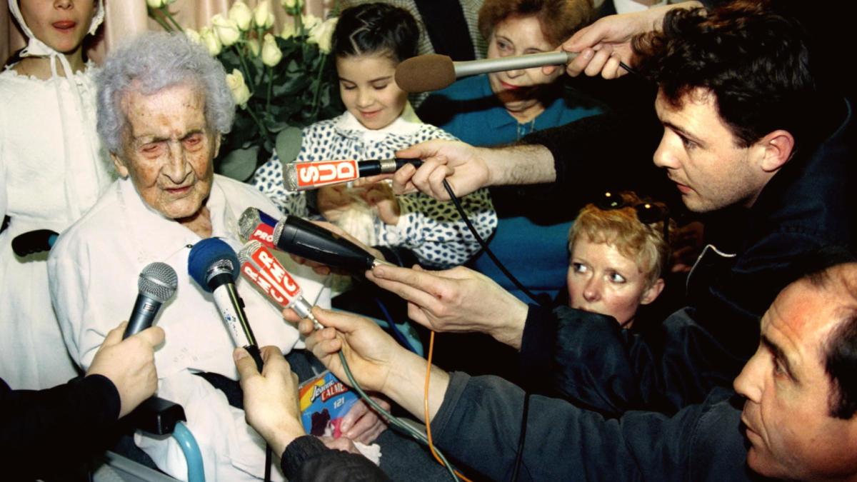 THE OLDEST PERSON IN THE WORLD, 2026, elderly woman in white sweater seated with members of press surrounding her with microphones